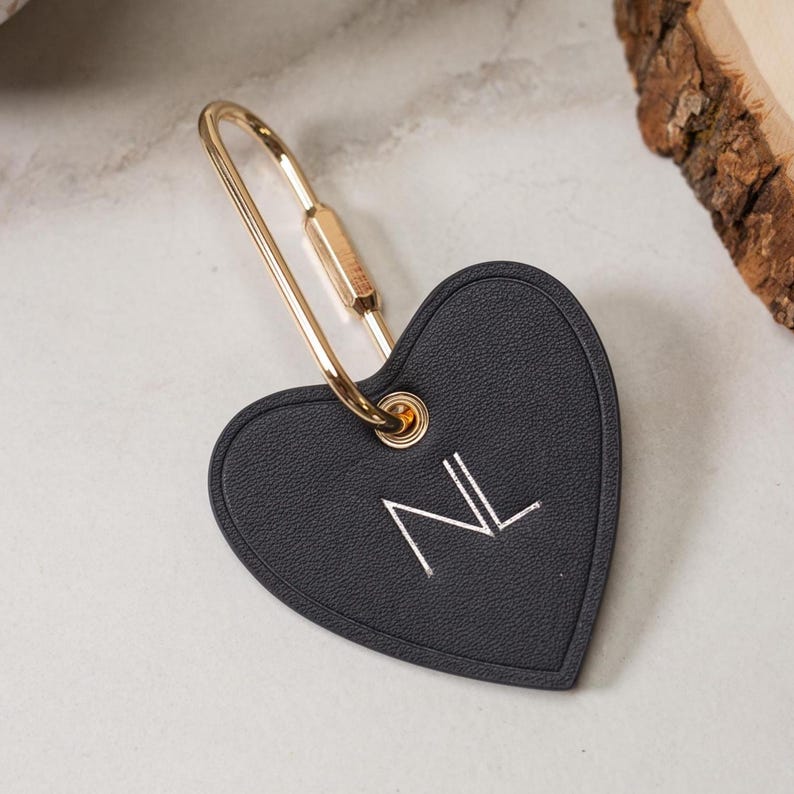 Puede incluir: Llavero negro en forma de coraz&oacute;n con cierre dorado. El llavero presenta las iniciales "NL" bordadas en blanco. El dise&ntilde;o es simple y elegante, adecuado para uso personal o como regalo.