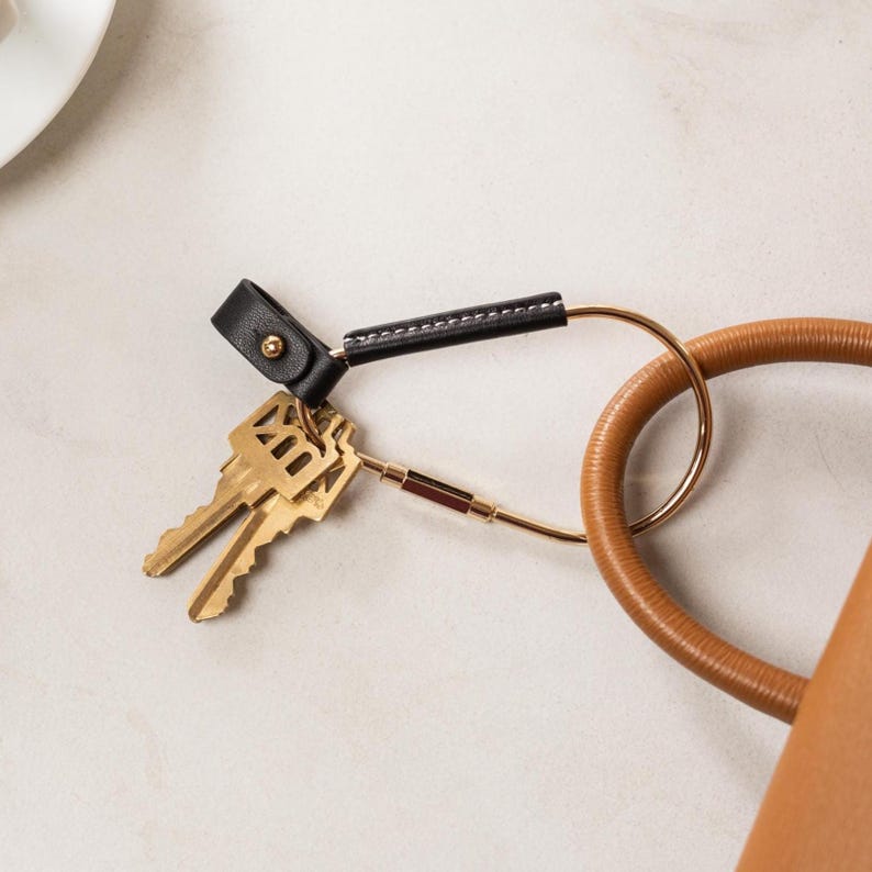 May include: A black leather key fob with gold-colored hardware and a gold key ring. The fob is attached to two gold keys. A brown leather handle is visible in the background.