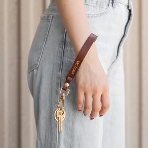 May include: A brown leather wrist strap with gold-colored hardware and a key. The strap is engraved with the name "VICTORIA" in gold lettering. The key has a gold-colored key ring.