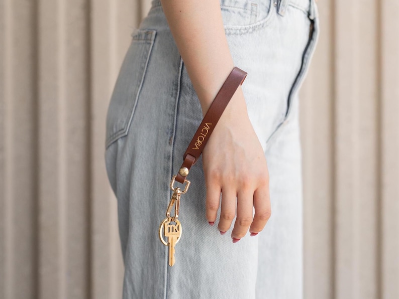 May include: A brown leather wrist strap with gold-colored hardware and a key. The strap is engraved with the name "VICTORIA" in gold lettering. The key has a gold-colored key ring.
