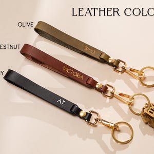May include: Leather keychains in olive, chestnut, and ebony. Each keychain features a gold-colored clasp and ring, with personalized initials or names. The text "LEATHER COLORS" is at the top.