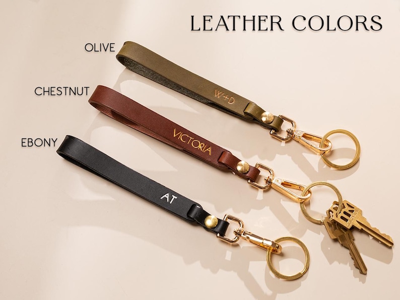 May include: Leather keychains in olive, chestnut, and ebony. Each keychain features a gold-colored clasp and ring, with personalized initials or names. The text "LEATHER COLORS" is at the top.