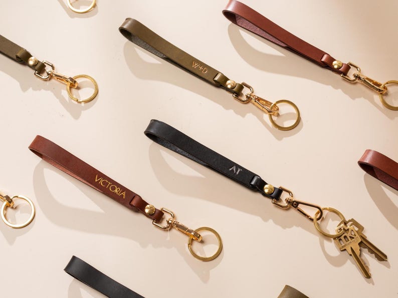 May include: Leather keychains in various colors, including brown, black, and olive green. Each keychain features a gold-colored clasp and ring, with some personalized with initials or names. Keys are attached to one keychain.