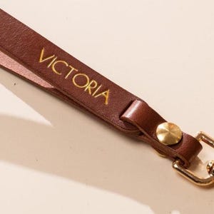 May include: A brown leather wrist strap with the name "VICTORIA" in gold lettering. The strap has a gold-colored clasp and a gold button. The strap is made of a smooth, durable leather material.