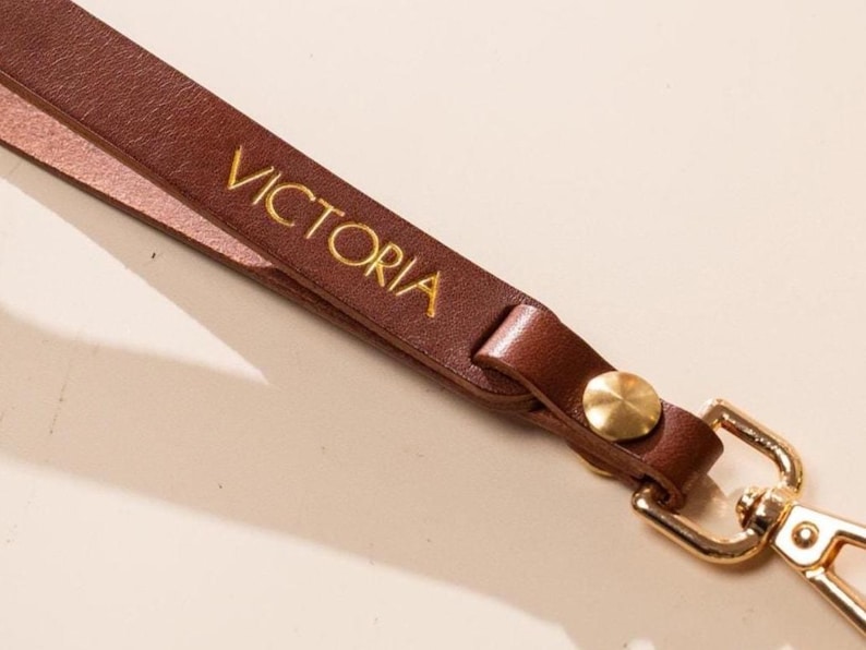 May include: A brown leather wrist strap with the name "VICTORIA" in gold lettering. The strap has a gold-colored clasp and a gold button. The strap is made of a smooth, durable leather material.