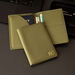 Puede incluir: Un portapasaportes de color verde oliva con las iniciales EC grabadas en oro en la cubierta frontal. El interior incluye un bolsillo para el pasaporte y ranuras para tarjetas, con un pasaporte parcialmente insertado en el lado izquierdo.