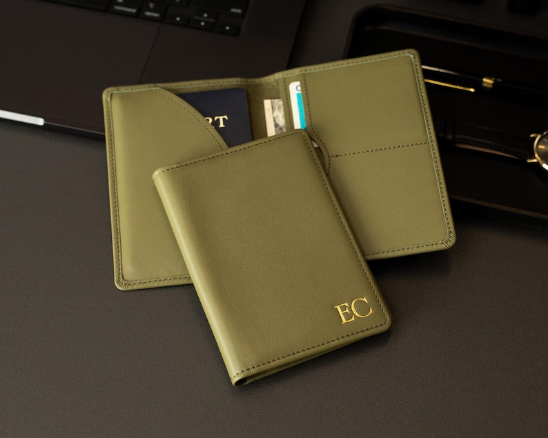 Puede incluir: Un portapasaportes de color verde oliva con las iniciales EC grabadas en oro en la cubierta frontal. El interior incluye un bolsillo para el pasaporte y ranuras para tarjetas, con un pasaporte parcialmente insertado en el lado izquierdo.