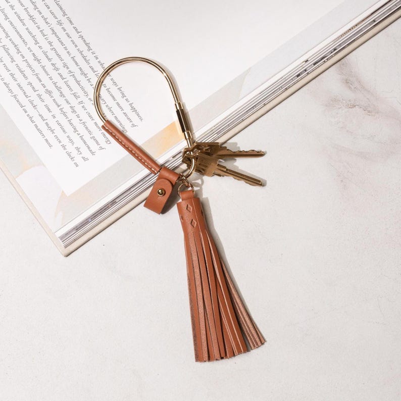 May include: A gold-toned metal keychain with a brown leather tassel and a small leather strap. The keychain holds several keys and is resting on a book. The tassel has a diamond pattern.
