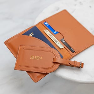 Puede incluir: Un portapasaporte de cuero color canela y una etiqueta de equipaje a juego con el nombre EILEEN grabado en oro. El portapasaporte abierto contiene un pasaporte azul, un billete de veinte d&oacute;lares y dos tarjetas de cr&eacute;dito en sus ranuras.