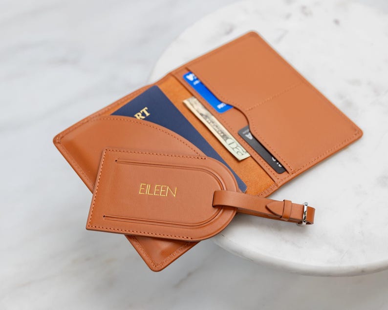Puede incluir: Un portapasaporte de cuero color canela y una etiqueta de equipaje a juego con el nombre EILEEN grabado en oro. El portapasaporte abierto contiene un pasaporte azul, un billete de veinte d&oacute;lares y dos tarjetas de cr&eacute;dito en sus ranuras.