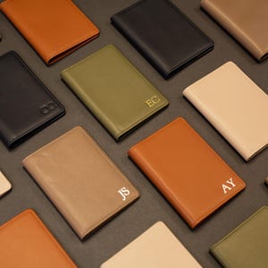 Puede incluir: Una colecci&oacute;n de fundas rectangulares para pasaporte en varios colores, incluyendo negro, tostado, verde oliva y crema. Varias de las cubiertas de aspecto cuero presentan monogramas dorados de dos letras como EC, ML, JS y AY, dispuestos en cuadr&iacute;cula.