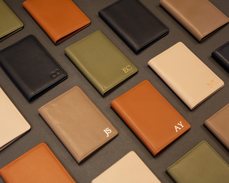 Puede incluir: Una colecci&oacute;n de fundas rectangulares para pasaporte en varios colores, incluyendo negro, tostado, verde oliva y crema. Varias de las cubiertas de aspecto cuero presentan monogramas dorados de dos letras como EC, ML, JS y AY, dispuestos en cuadr&iacute;cula.