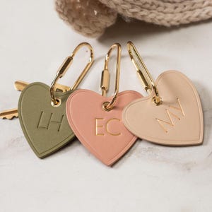 Puede incluir: Tres llaveros en forma de coraz&oacute;n en verde oliva, rosa y beige, cada uno con herrajes dorados. Los llaveros tienen iniciales grabadas: "LH", "EC" y "MY".