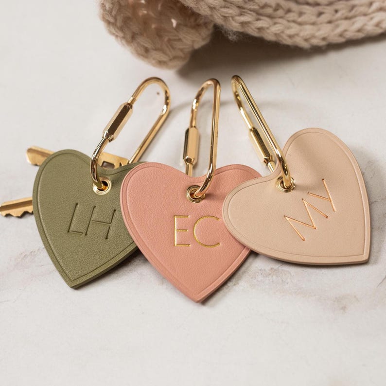 Puede incluir: Tres llaveros en forma de coraz&oacute;n en verde oliva, rosa y beige, cada uno con herrajes dorados. Los llaveros tienen iniciales grabadas: "LH", "EC" y "MY".