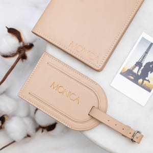 Puede incluir: Un portapasaporte de cuero beige y una etiqueta de equipaje a juego, ambos grabados con el nombre MONICA en letras doradas. La etiqueta de equipaje cuenta con una correa de hebilla. Una peque&ntilde;a foto rectangular muestra la silueta de una pareja cerca de la Torre Eiffel.