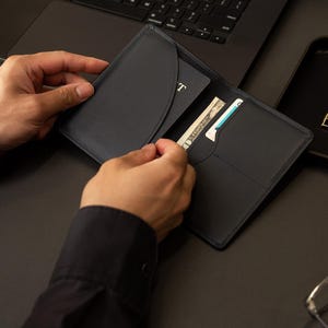 Puede incluir: Un portapasaportes de cuero azul marino oscuro con un bolsillo interior curvo para el pasaporte, una ranura vertical para billetes y un compartimento adicional para tarjetas. El dise&ntilde;o incluye costuras tonales y un acabado mate liso.