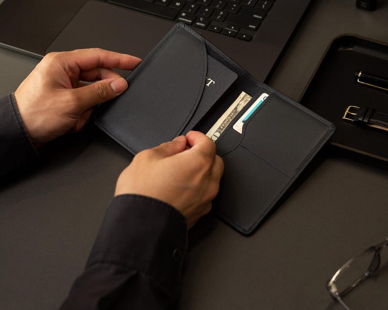 Puede incluir: Un portapasaportes de cuero azul marino oscuro con un bolsillo interior curvo para el pasaporte, una ranura vertical para billetes y un compartimento adicional para tarjetas. El dise&ntilde;o incluye costuras tonales y un acabado mate liso.