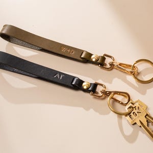 May include: Two leather keychains, one olive green and one black, with gold-colored hardware. The olive green keychain has the initials "W+D" and the black keychain has "AT". Keys are attached to one keychain.