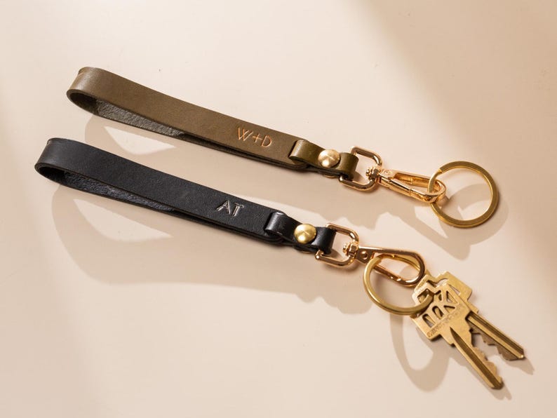 May include: Two leather keychains, one olive green and one black, with gold-colored hardware. The olive green keychain has the initials "W+D" and the black keychain has "AT". Keys are attached to one keychain.