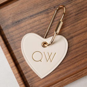Puede incluir: Llavero en forma de coraz&oacute;n crema con herrajes dorados. El llavero presenta las letras doradas "QW". El llavero est&aacute; sobre una superficie de madera.