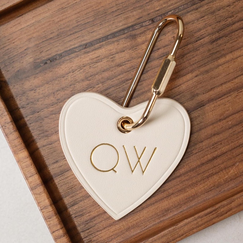 Puede incluir: Llavero en forma de coraz&oacute;n crema con herrajes dorados. El llavero presenta las letras doradas "QW". El llavero est&aacute; sobre una superficie de madera.