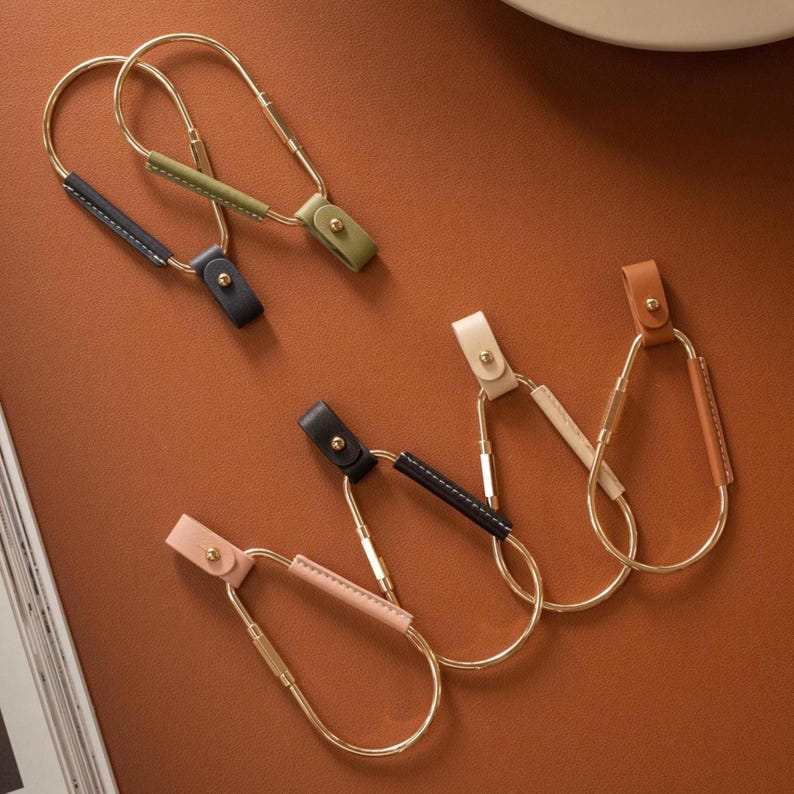 May include: A collection of gold-toned metal keychains with leather accents in various colors, including black, green, pink, beige, and brown. Each keychain features a loop design with a leather strap and a small button closure. The keychains are arranged on a brown surface.
