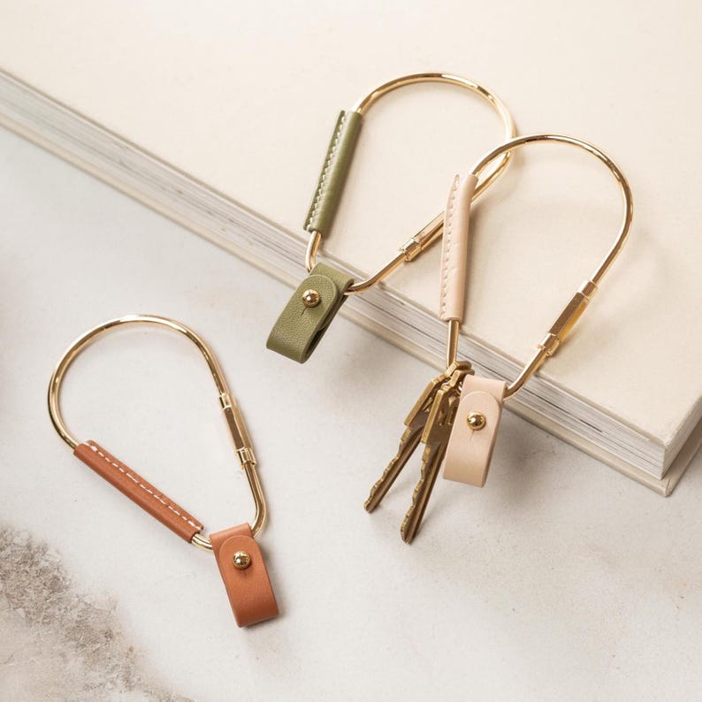 May include: Three keychains with gold-colored metal arches and leather accents in olive green, tan, and light pink. The keychains have a simple, modern design, with a leather strap and a gold-colored metal clasp. One keychain holds keys.