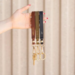 May include: Three leather wrist keychains in brown, olive green, and navy blue, each with gold hardware and personalized text. The keychains are held up against a neutral background.