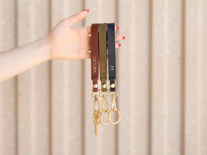 May include: Three leather wrist keychains in brown, olive green, and navy blue, each with gold hardware and personalized text. The keychains are held up against a neutral background.