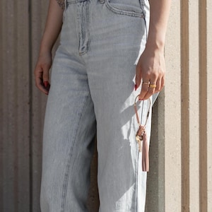 May include: Light wash denim jeans with a high waist. A person is holding a leather key fob with keys and a pink tassel. The person is wearing gold rings and has red nail polish.