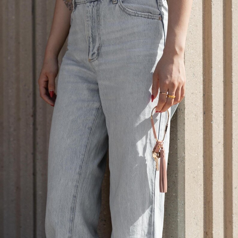 May include: Light wash denim jeans with a high waist. A person is holding a leather key fob with keys and a pink tassel. The person is wearing gold rings and has red nail polish.