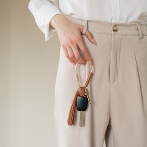 May include: A gold-toned keychain with a brown leather strap and tassel, holding a car key and fob. The keychain is held against a background of beige pants and a white shirt.