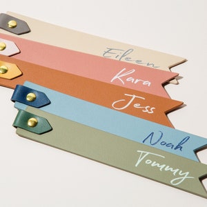 May include: Six colorful leather bookmarks with gold metal accents and names written in white. The bookmarks are arranged in a fan shape with the names "Eileen", "Kara", "Jess", "Noah", and "Tommy" visible.