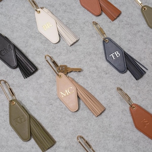 May include: Selection of personalised keyrings in various colours, including black, white, tan, and olive green. Each keyring features a tassel and gold-coloured hardware. The keyrings are engraved with initials.