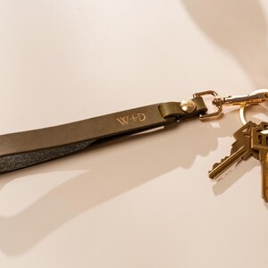 May include: Olive green leather keychain with gold-colored hardware and two keys. The keychain strap is embossed with the initials "W+D". The keys are attached to a gold-colored split ring.