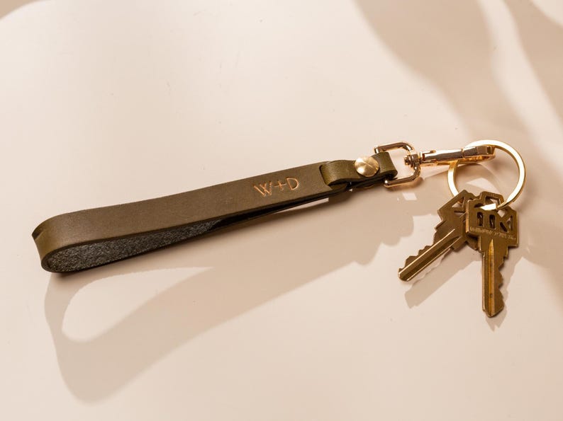 May include: Olive green leather keychain with gold-colored hardware and two keys. The keychain strap is embossed with the initials "W+D". The keys are attached to a gold-colored split ring.
