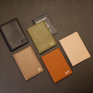 Puede incluir: Una colecci&oacute;n de cinco fundas rectangulares para pasaporte en negro, verde oliva, beige, tostado y marr&oacute;n, cada una con monogramas de dos letras en relieve. Una funda est&aacute; abierta y muestra la palabra PASSPORT en la cubierta interior.