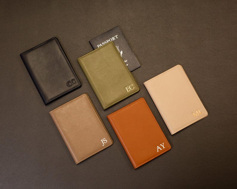 Puede incluir: Una colecci&oacute;n de cinco fundas rectangulares para pasaporte en negro, verde oliva, beige, tostado y marr&oacute;n, cada una con monogramas de dos letras en relieve. Una funda est&aacute; abierta y muestra la palabra PASSPORT en la cubierta interior.