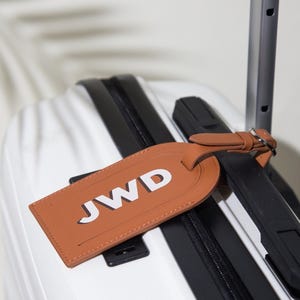 May include: A tan luggage tag with the letters JWD in white, attached by a matching strap to the handle of a white suitcase with black accents. The rectangular tag features rounded corners and visible stitching along the edges.