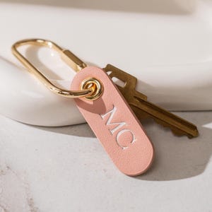 May include: A close-up of a pink leather keychain with the initials "MC" in white. The keychain is attached to a gold-colored clasp and a brass key. The background is a blurred white surface.
