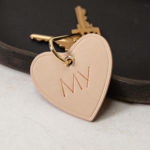 Puede incluir: Un llavero de cuero beige claro en forma de coraz&oacute;n con las letras "MY" grabadas y un anillo de metal dorado. Una llave dorada es visible en el fondo. El accesorio es personalizado.