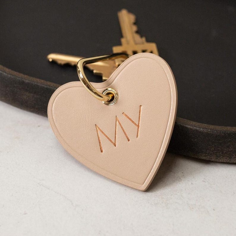 Puede incluir: Un llavero de cuero beige claro en forma de coraz&oacute;n con las letras "MY" grabadas y un anillo de metal dorado. Una llave dorada es visible en el fondo. El accesorio es personalizado.