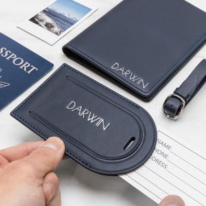 Puede incluir: Un portapasaportes de cuero azul marino y una etiqueta de equipaje a juego, ambos personalizados con el nombre DARWIN en letras blancas. La etiqueta de equipaje cuenta con una tarjeta de informaci&oacute;n blanca extra&iacute;ble con campos para nombre, tel&eacute;fono y direcci&oacute;n.