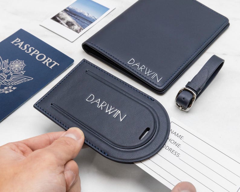 Puede incluir: Un portapasaportes de cuero azul marino y una etiqueta de equipaje a juego, ambos personalizados con el nombre DARWIN en letras blancas. La etiqueta de equipaje cuenta con una tarjeta de informaci&oacute;n blanca extra&iacute;ble con campos para nombre, tel&eacute;fono y direcci&oacute;n.