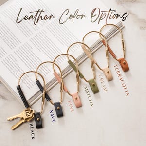 May include: A selection of leather keychains in various colors, including black, navy, pink, matcha, cream, and terracotta. Each keychain features a gold-colored metal loop and a leather strap with a snap closure. The text "Leather Color Options" is displayed.