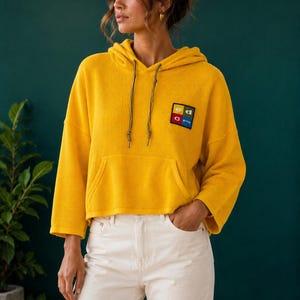 Può includere: Una felpa con cappuccio in maglia a coste giallo brillante, caratterizzata da una lunghezza corta, maniche a tre quarti e una tasca a marsupio frontale. Una toppa quadrata sul petto mostra il testo "ed" sopra "c group" in lettere bianche su quattro quadrati colorati.