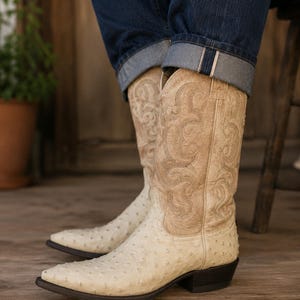 Puede incluir: Un par de botas vaqueras de color crema con un patrón texturizado en el pie y una caña de color tostado claro adornada con intrincados diseños de volutas cosidas en tonos a juego. Las botas tienen punta afilada y un tacón apilado marrón oscuro.