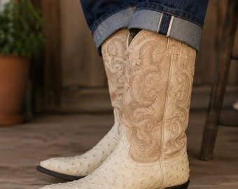 May include: A pair of cream-colored cowboy boots featuring a textured, quill-like pattern on the foot and a lighter tan shaft adorned with intricate, tonal stitched scrollwork designs. The boots have a pointed toe and a dark brown stacked heel.