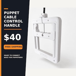 May include: A white rectangular puppet cable control handle featuring a central sliding mechanism and a vertical rod attachment point. The frame has a structured design with a grip area and the text LUMINOUS ENCHANTMENTS embossed on the bottom.