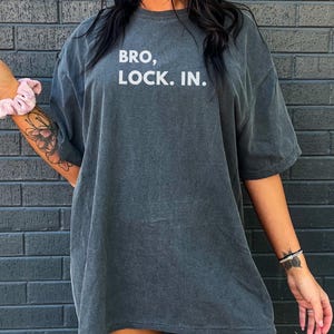 May include: A dark grey oversized t-shirt dress with the white text "BRO, LOCK. IN.". The model is holding a beverage in a clear cup. The shirt has a washed-out appearance.